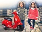Dve deti pózujú vedľa červeného detského skútra Vespa.