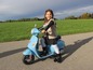 Dievča jazdí na modrej elektrickej motorke, podobnej Vespa.