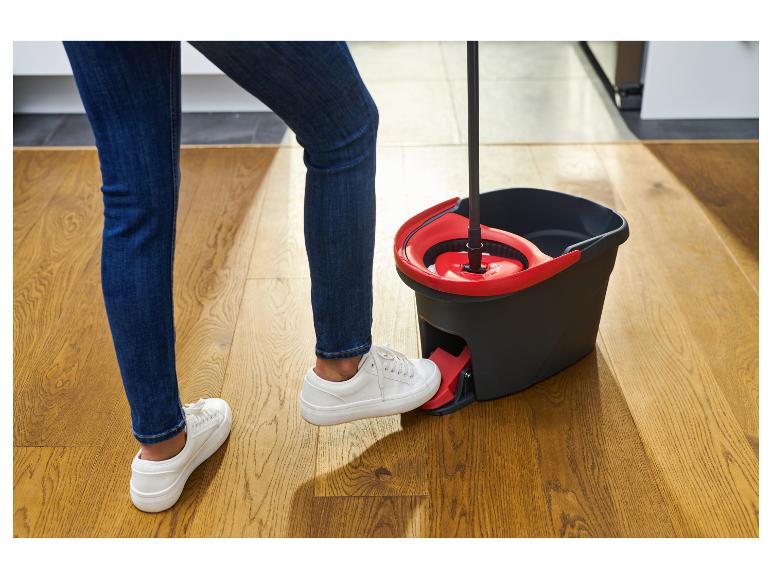 Osoba v džínsoch a bielych topánkach používa mop s vedrom s pedálom.