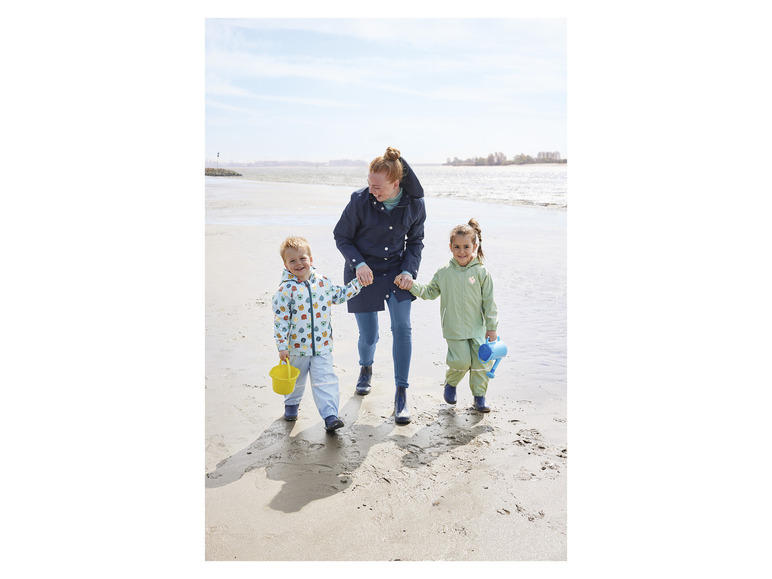 Rodina kráča po pláži v dažďovom oblečení, dieťa drží vedro.