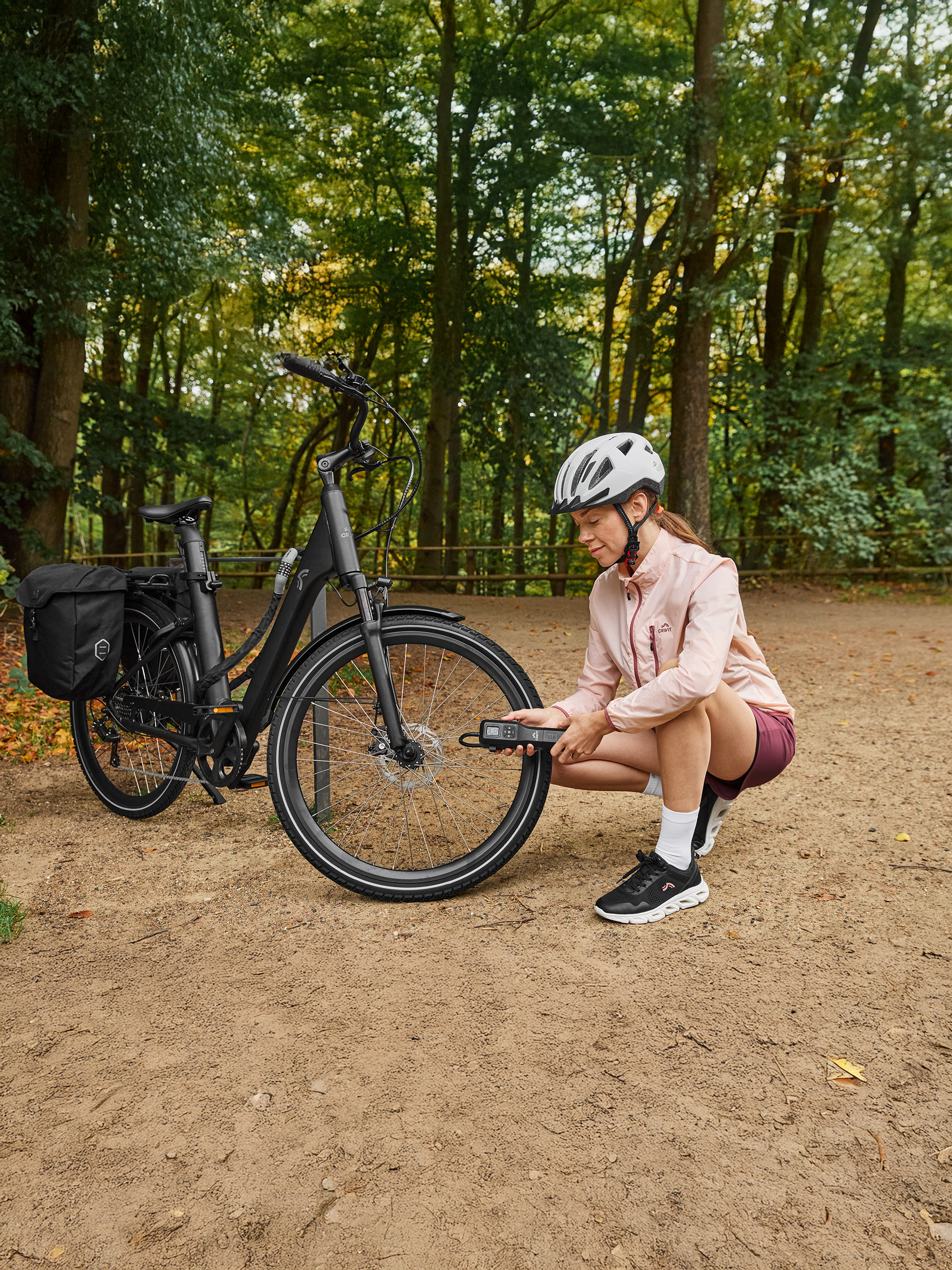Žena v športovom oblečení Crivit kontroluje tlak v pneumatikách e-bicykla prenosnou pumpou v lese.