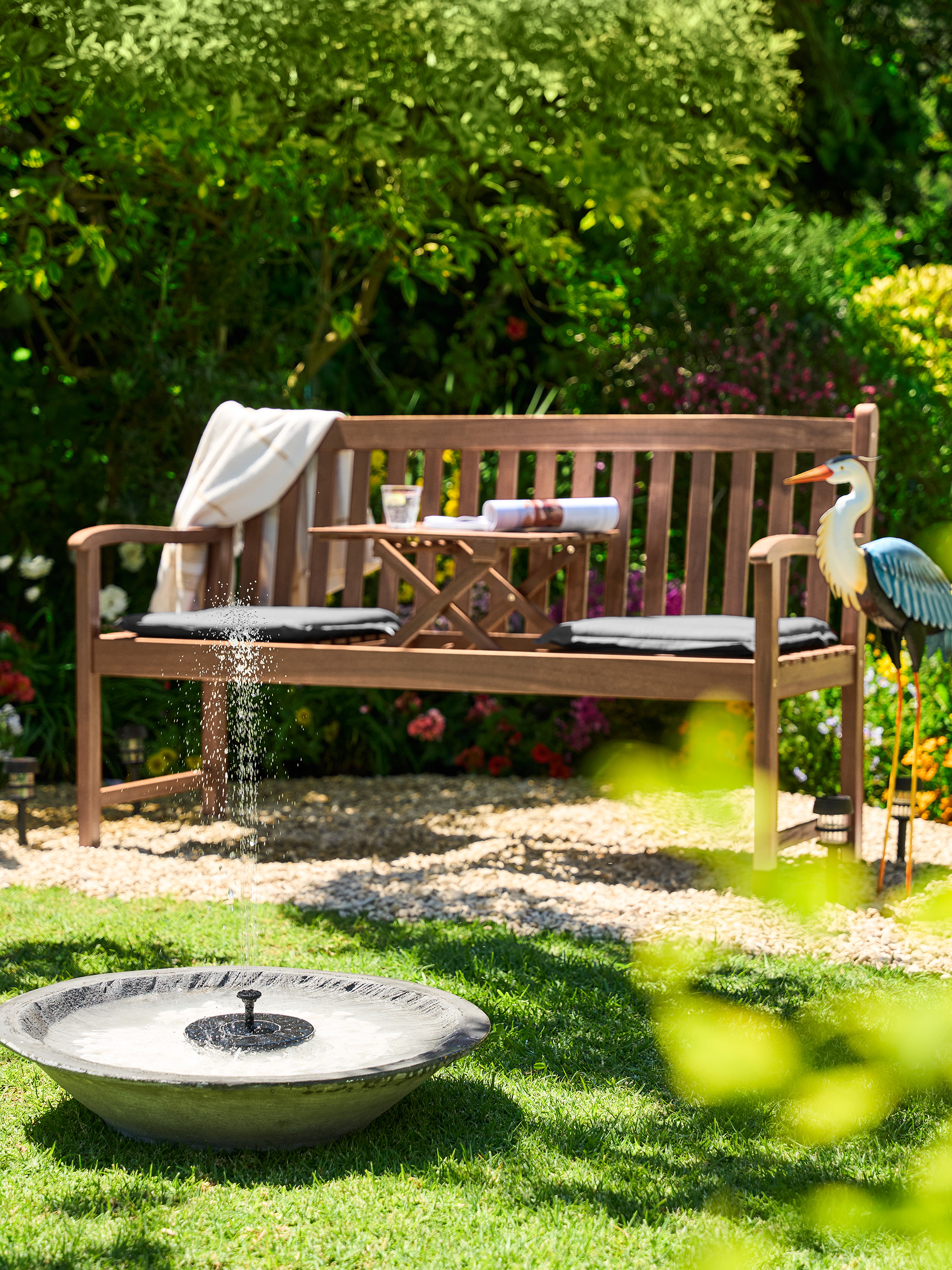 Drevená záhradná lavička s malým stolíkom, vankúšmi a dekoratívnou volavkou v slnečnej záhrade.