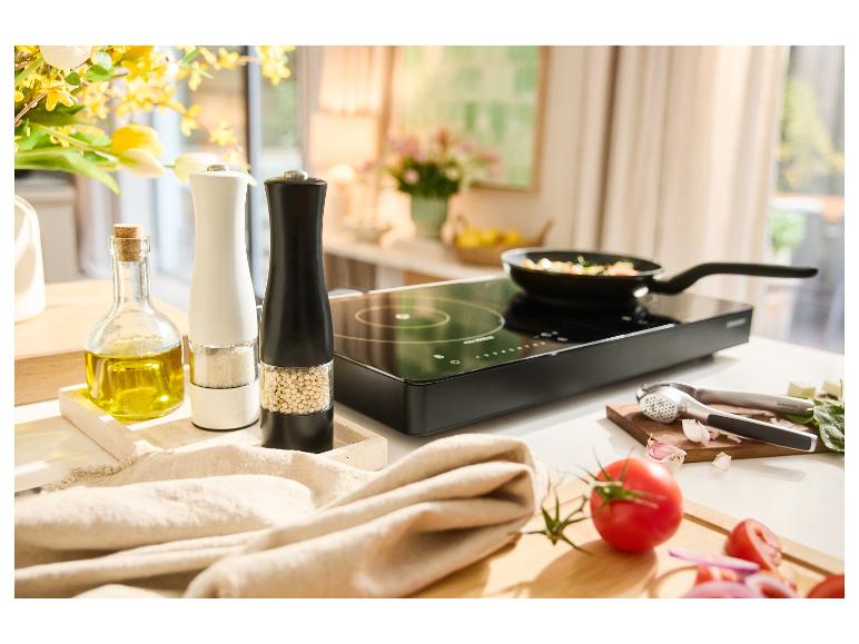 Indukčná varná doska s panvicou, koreničkami, olivovým olejom a čerstvou zeleninou na kuchynskej linke.