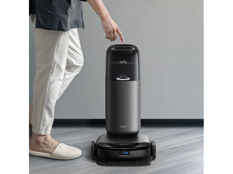 Osoba stláča robotický vysávač so samočistiacou stanicou na drevenej podlahe.