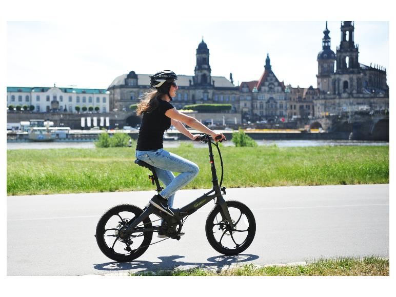 Žena jazdí na elektrobicykli Sachsengrad po nábreží s panorámou mesta v pozadí.