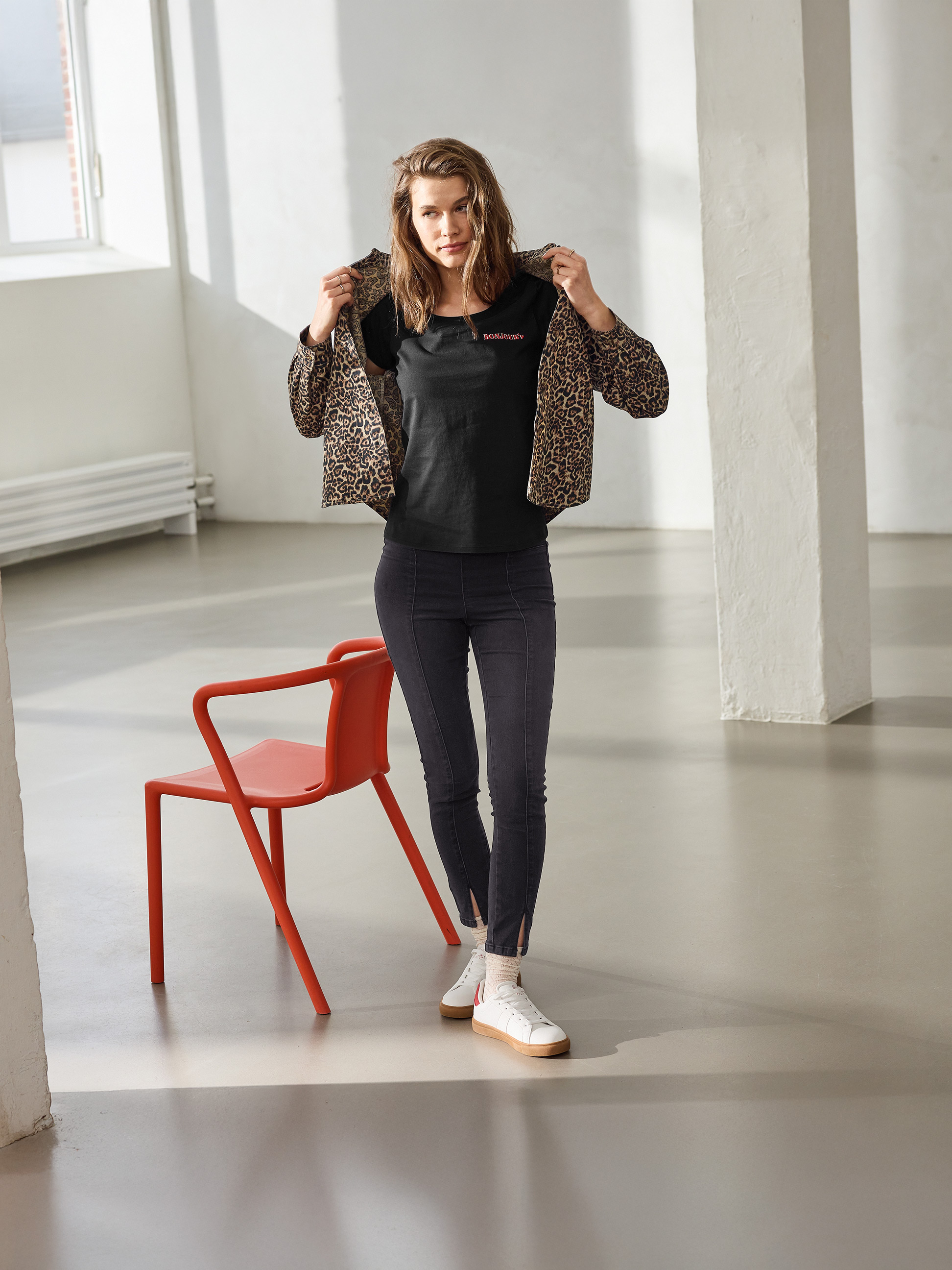 Žena v leopardej bunde, čiernom tričku 'BONJOUR' a čiernych úzkych džínsoch, pózujúca vedľa červenej stoličky.