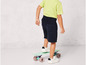 Dieťa v zelenom tričku a modrých šortkách stojí na skateboardu.