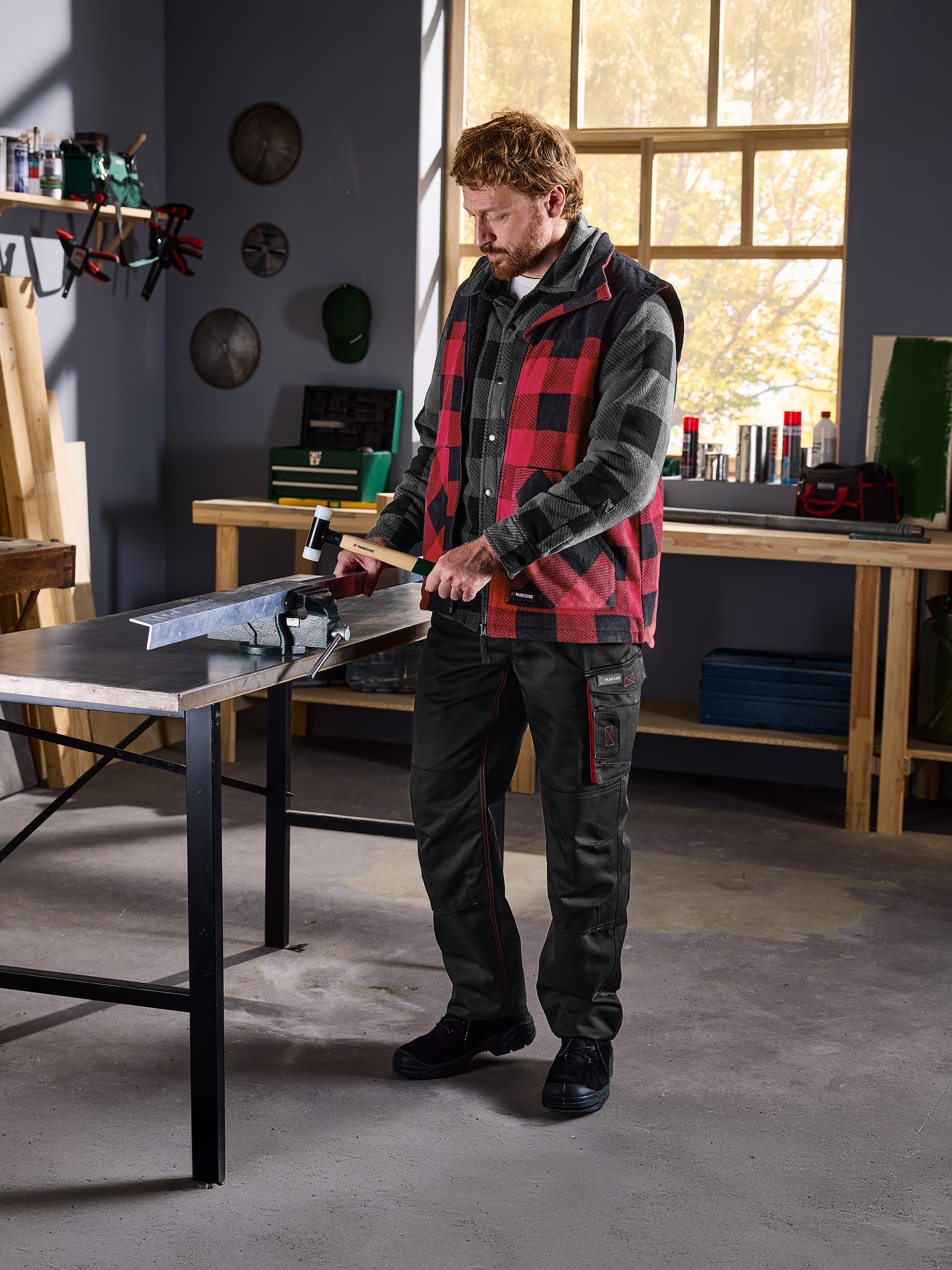 Muž v kockovanej košeli a veste Parkside pracuje s kovom vo zveráku v dielni.