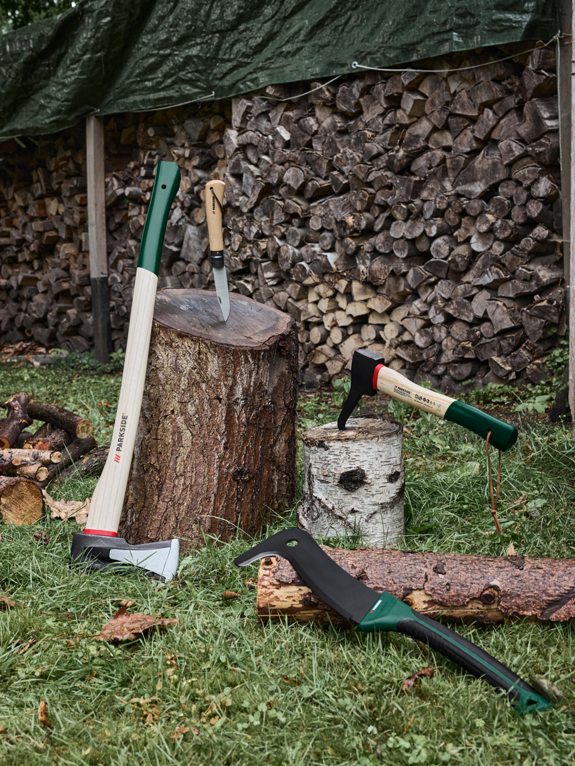 Sekery, nôž a mačeta na tráve s naskladaným drevom v pozadí.