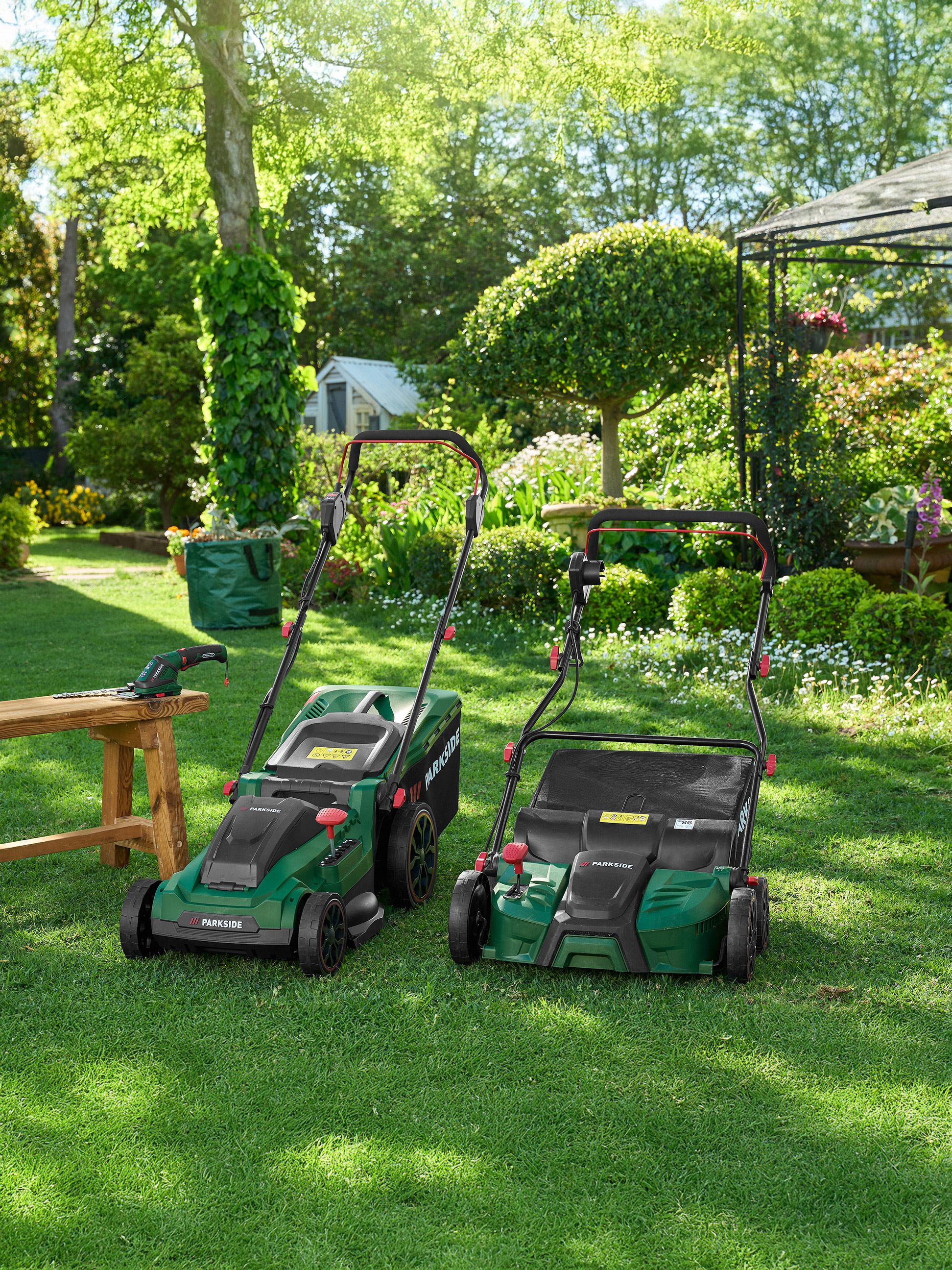 Dve zelené kosačky Parkside a záhradné náradie na trávniku v záhrade.