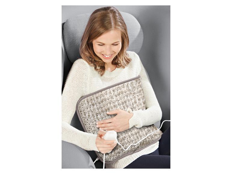 Žena v kresle sa usmieva, zatiaľ čo drží béžovú elektrickú vyhrievaciu podložku na kolenách.