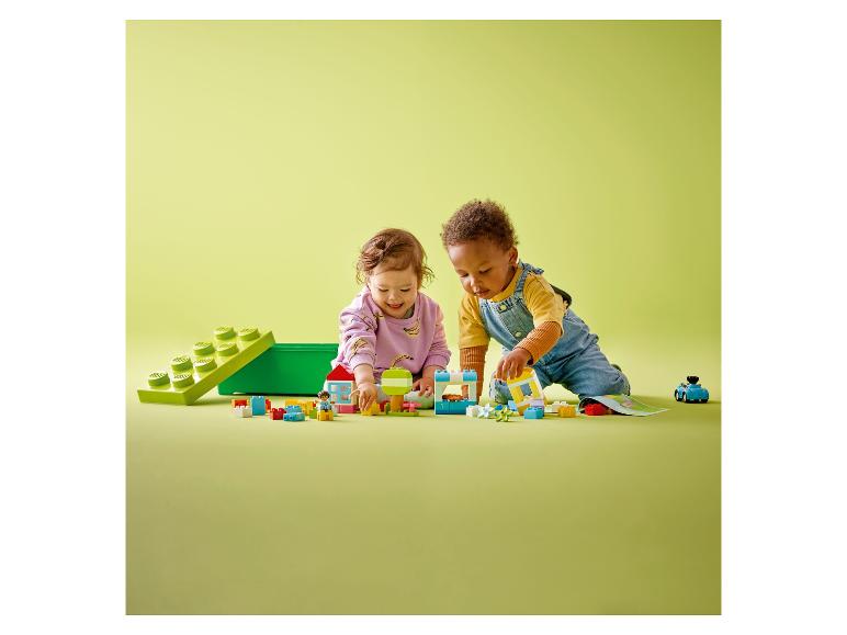 Dve batoľatá sa hrajú s farebnými kockami a figúrkami LEGO DUPLO na zelenom povrchu.