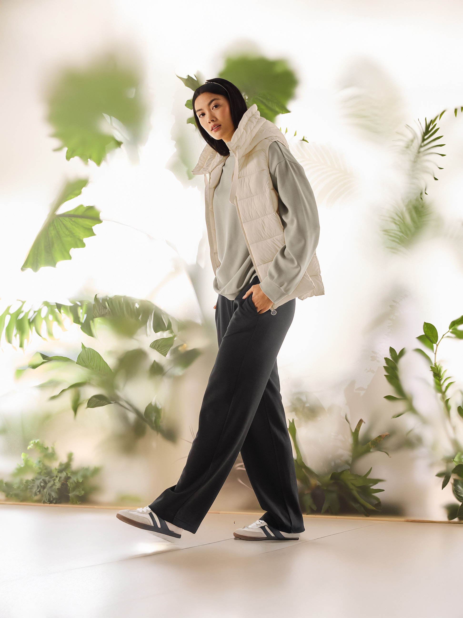 Žena v béžovej prešívanej veste, svetlozelenej mikine a čiernych nohaviciach kráča.