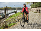 Žena v prilbe a červenej bunde jazdí na čiernom elektrobicykli po dláždenej ceste pri rieke.