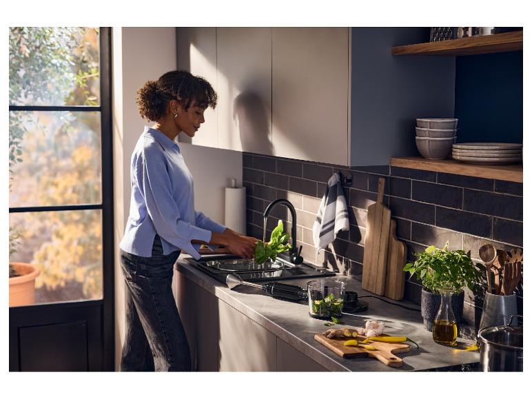 Žena umýva špenát v modernej kuchyni s ponorným mixérom a prísadami na pulte.