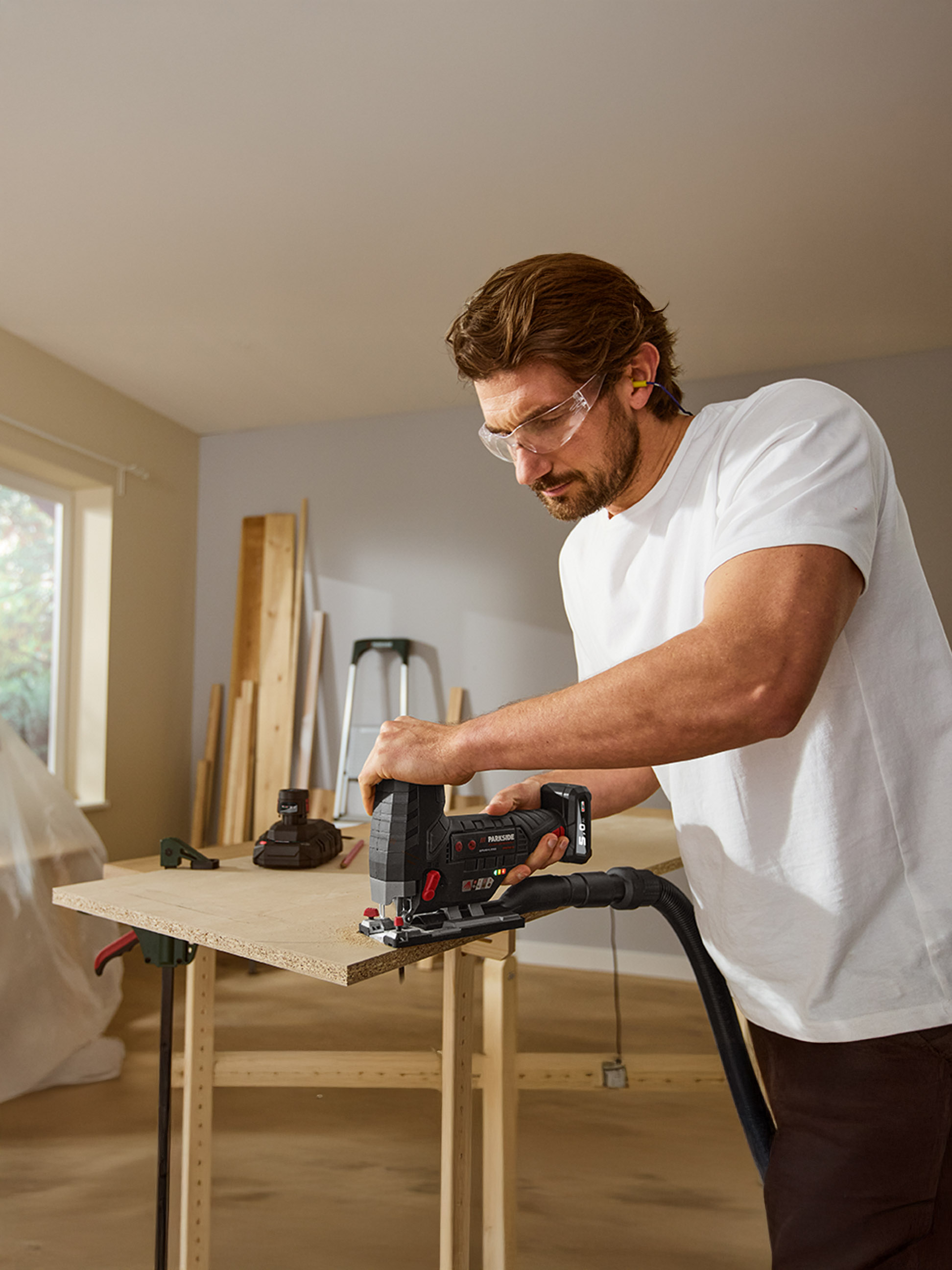 Muž v ochranných okuliaroch a štuple do uší reže drevo akumulátorovou priamočiarou pílou.