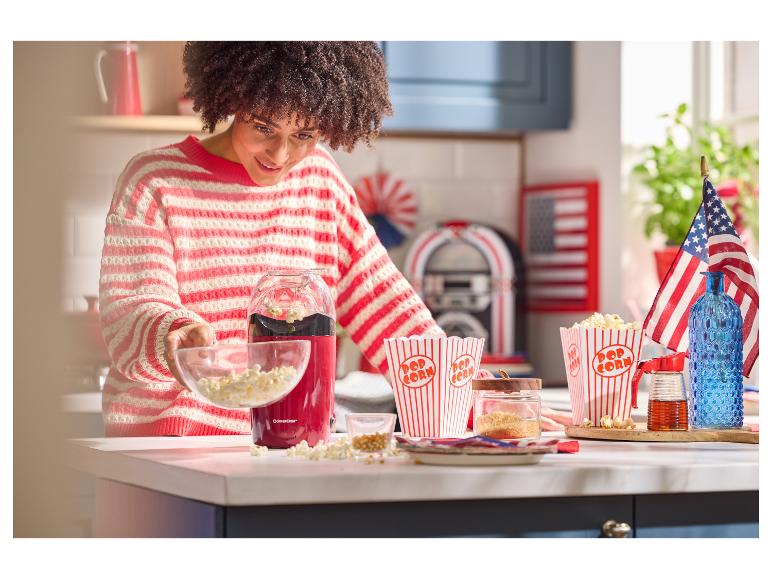Žena sype popcorn z červeného popcornovača do misky, s krabicami na popcorn a americkými vlajkami.
