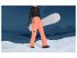 Žena v oranžových lyžiarskych nohaviciach s snowboardom a horským prostredím v pozadí.