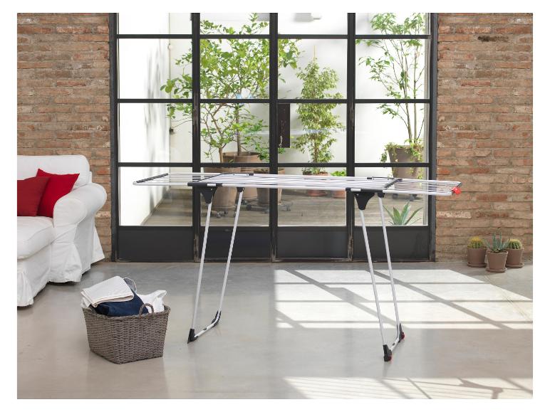 Biely sušiak na bielizeň v modernom interiéri s tehlovou stenou a rastlinami.