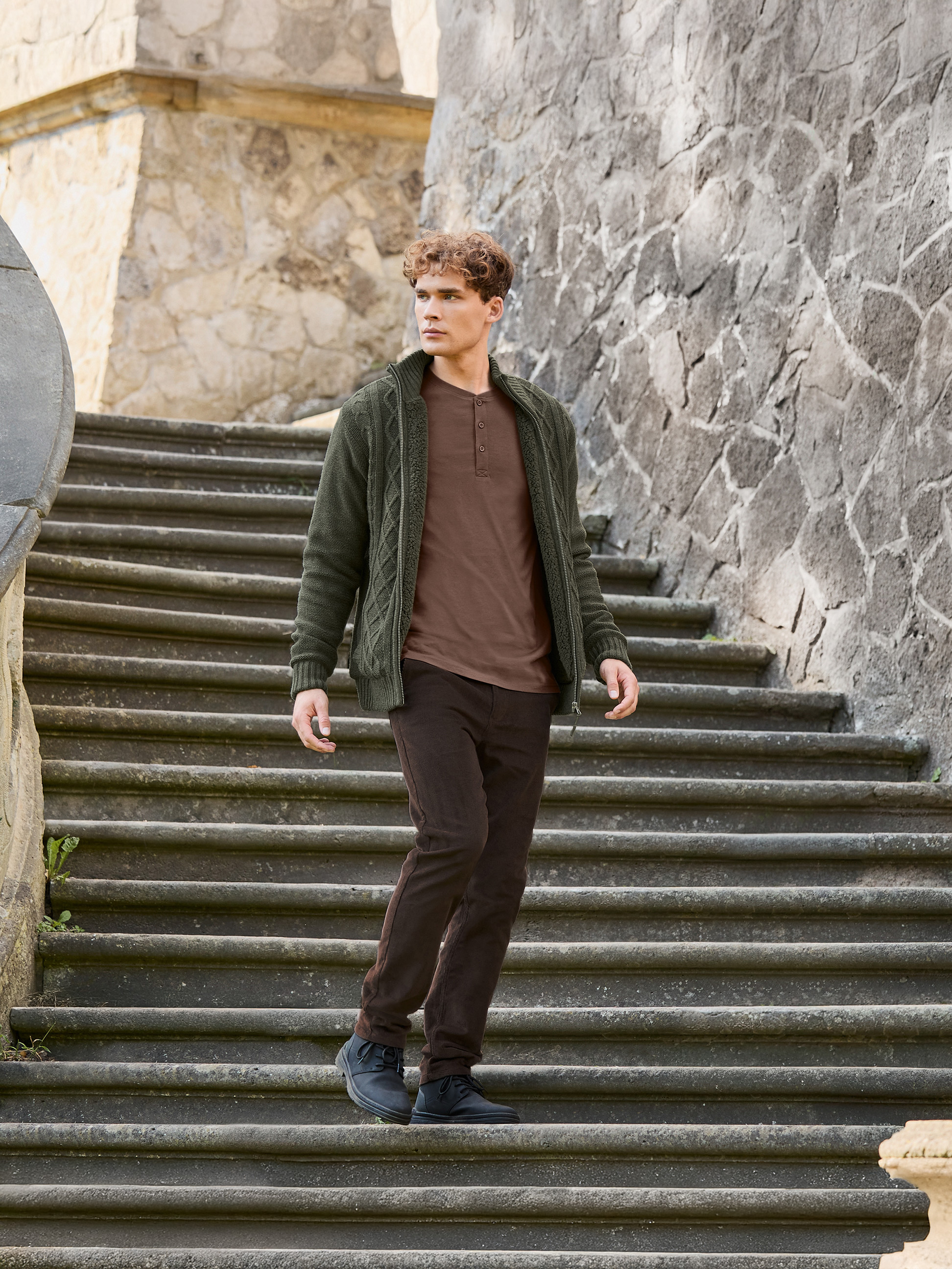 Muž v zelenom pletenom kabáte, hnedom tričku a hnedých nohaviciach schádza po kamenných schodoch.