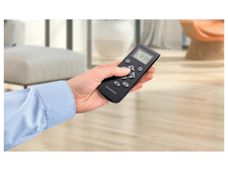 Ruka drží čierny diaľkový ovládač s digitálnym displejom zobrazujúcim 12:33, v svetlej miestnosti.