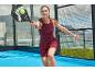 Žena v bordových športových šatách hrá padel s raketou a loptičkou.