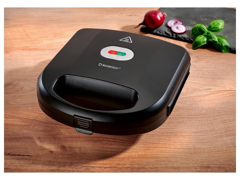 Čierny sendvičovač SilverCrest na drevenom stole s cibuľou a bazalkou.