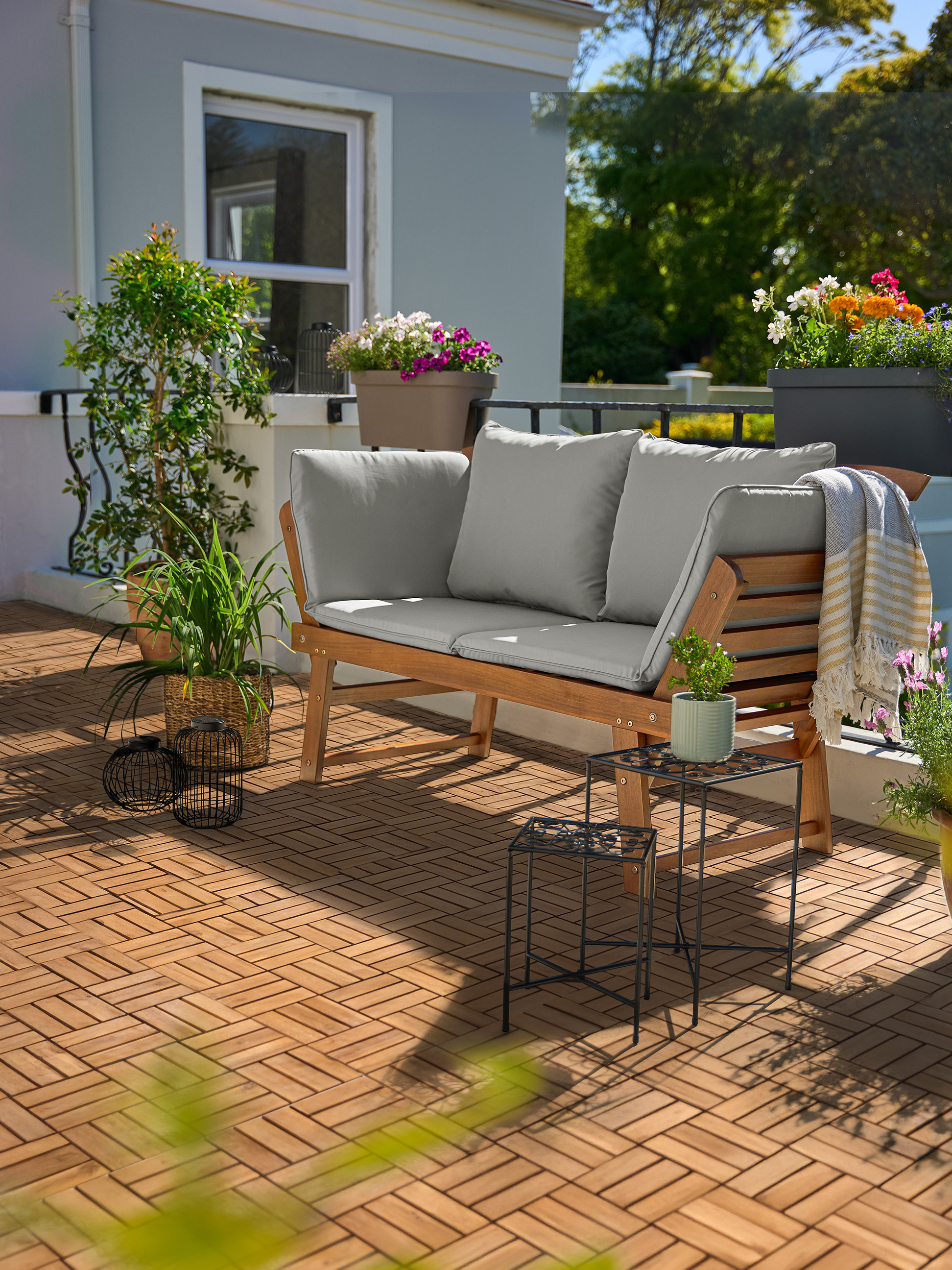 Drevená záhradná pohovka so sivými vankúšmi na balkóne s drevenými dlaždicami a rastlinami