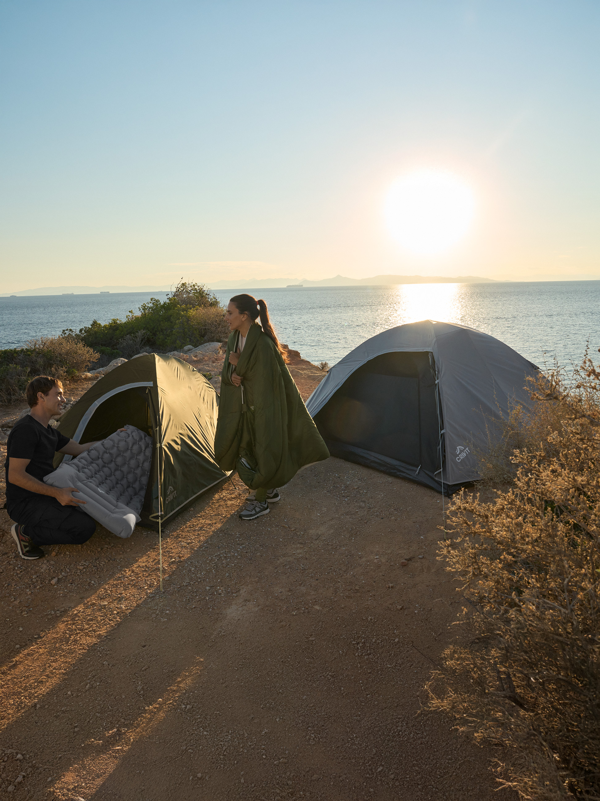Dvaja ľudia kempujú pri mori s dvoma stanmi Crivit a nafukovacím matracom.