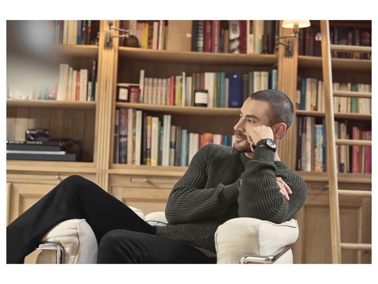 Muž v tmavozelenom pletenom svetri a čiernych nohaviciach sedí v kresle pred knižnicou.