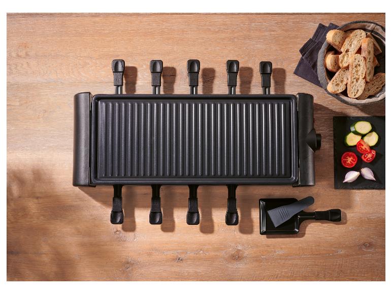 Raclette gril so šiestimi panvicami, chlebom a nakrájanou zeleninou na drevenom stole.