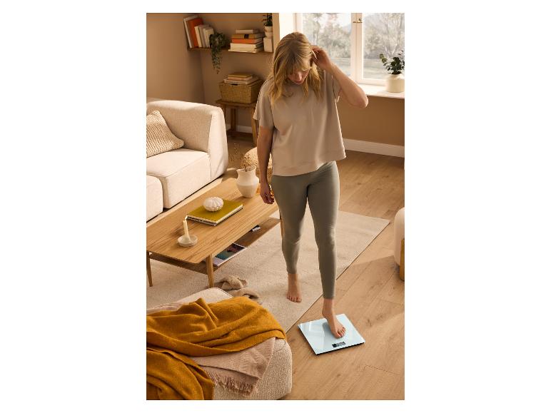 Mladá žena v tričku a legínach, vážiaca sa na digitálnej kúpeľňovej váhe.