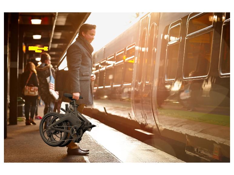 Muž v kabáte drží skladací bicykel na nástupišti vedľa vlaku.