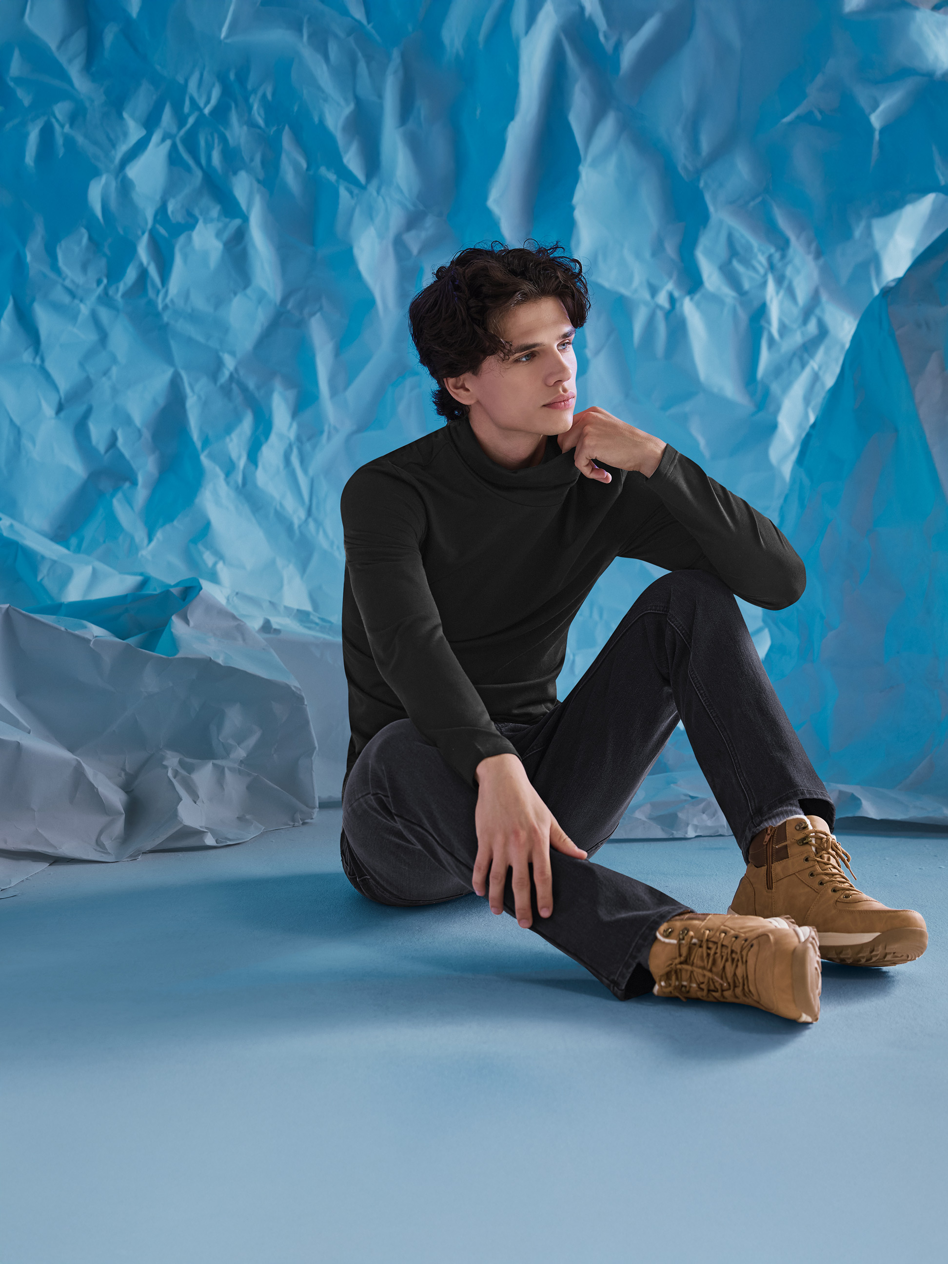 Muž v čiernom roláku, džínsoch a hnedých čižmách, sediaci na modrom pozadí.