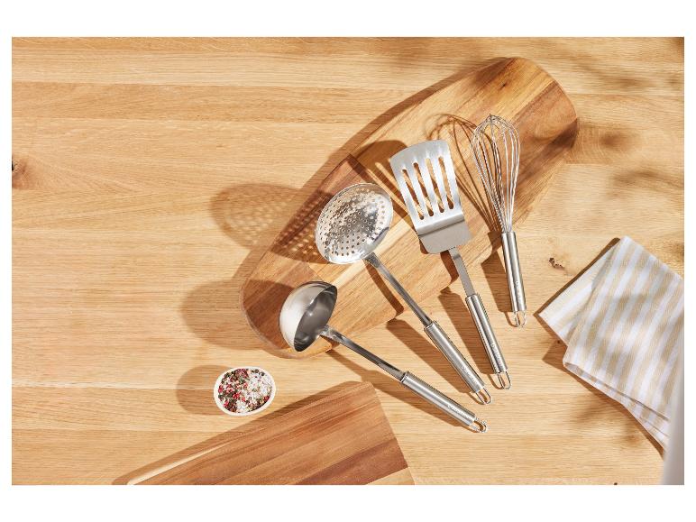 Nerezové kuchynské náčinie vrátane špachtle, naberačky a metličky na drevenej doske na krájanie.