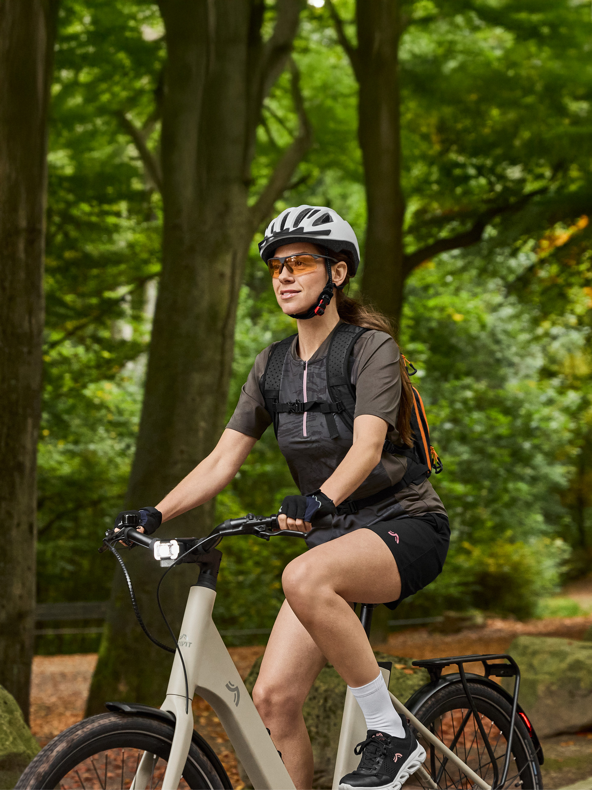 Žena na elektrobicykli s prilbou, slnečnými okuliarmi a batohom v lese.
