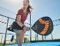 Žena hrá padel s čiernou a oranžovou padelovou raketou na modrom kurte.