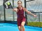 Žena v bordových športových šatách hrá padel s čiernou raketou a žltou loptičkou.