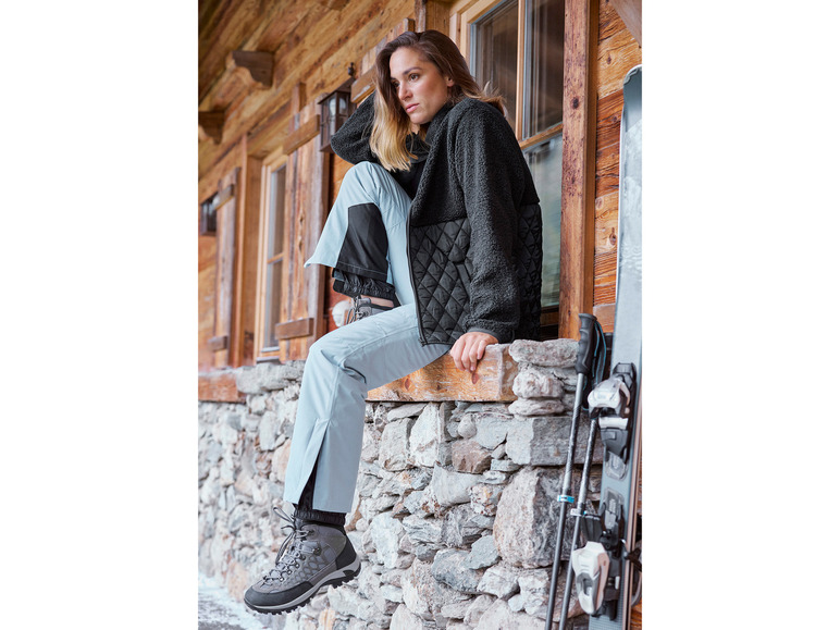 Žena v čiernej fleecovej bunde a modrých lyžiarskych nohaviciach s lyžami a palicami na pozadí.