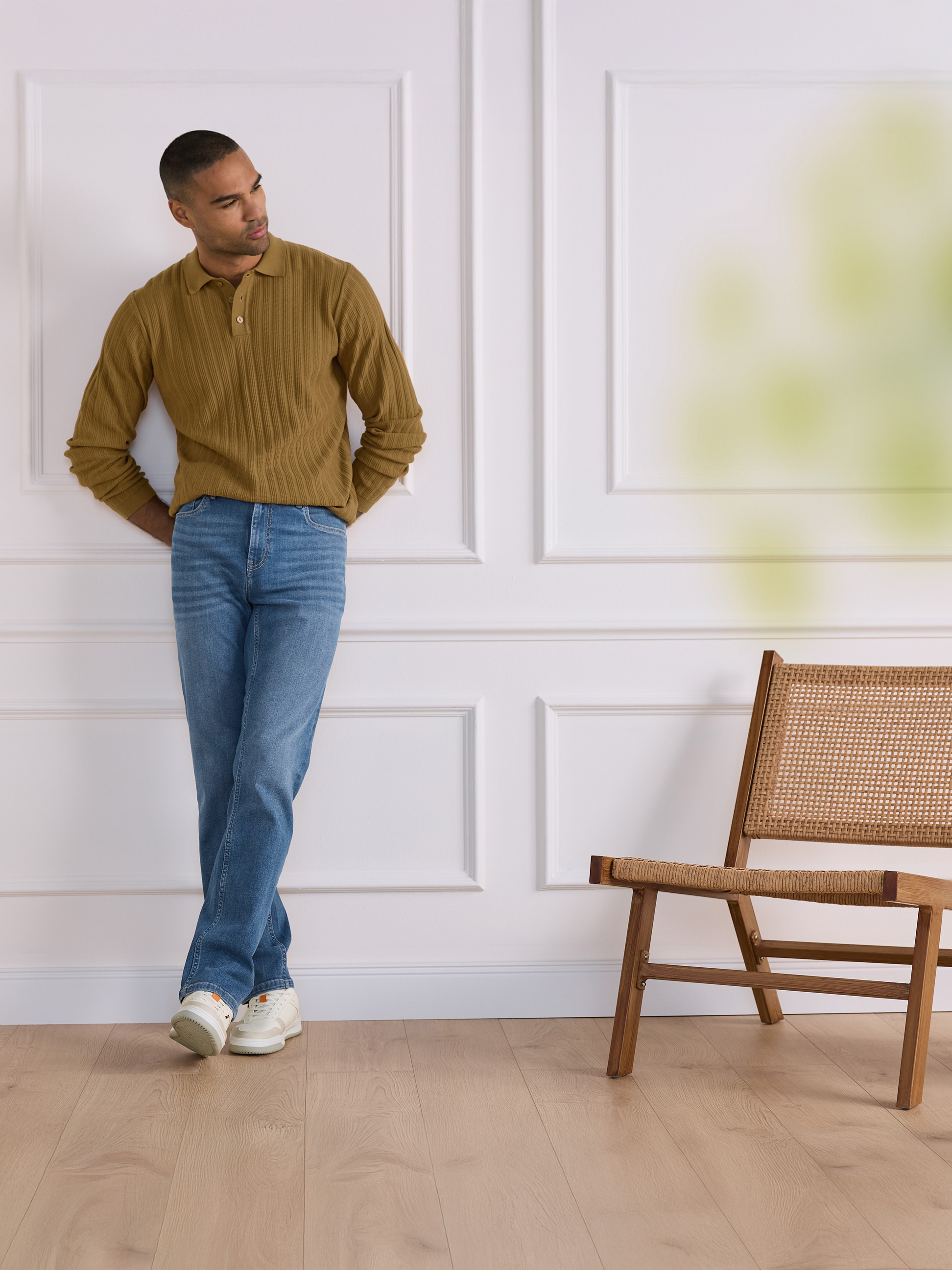 Muž v hnedom rebrovanom polotričku a džínsoch sa opiera o bielu stenu s drevenou stoličkou.