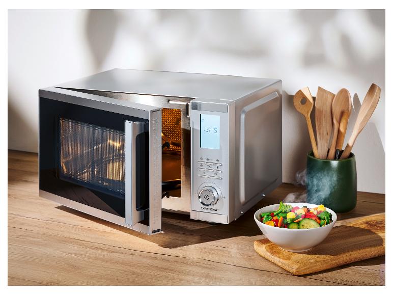 Strieborná mikrovlnná rúra s otvorenými dvierkami a miskou zeleniny na drevenej doske.