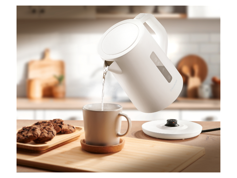 Biela elektrická kanvica SilverCrest naleje vodu do hrnčeka na drevenom stole.
