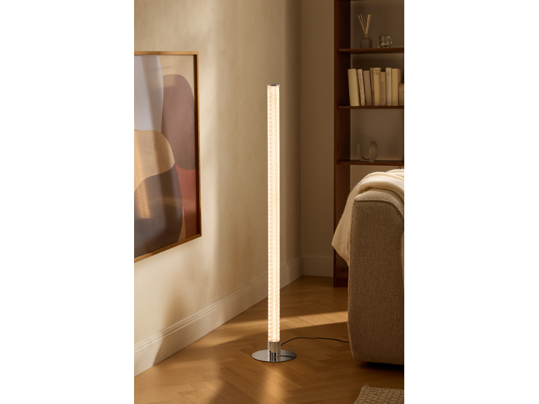 Valcová stojaca lampa s chrómovou základňou osvetľuje modernú obývačku s knižnicou a pohovkou.