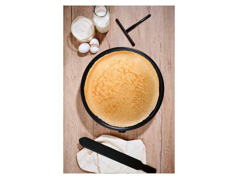 Pohľad zhora na palacinkovač so zlatou palacinkou, obklopený múkou, mliekom a vajcami.