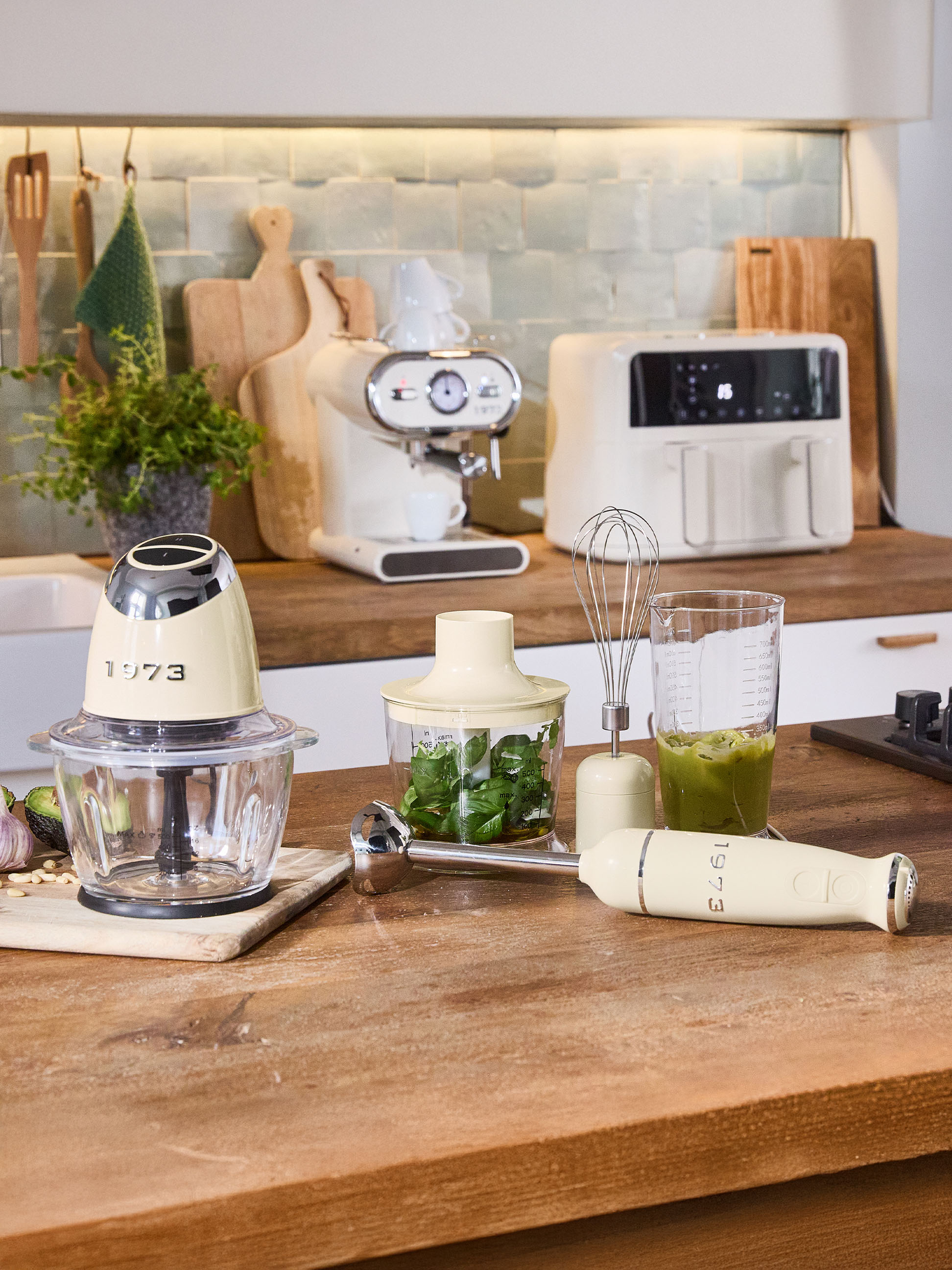 Krémové kuchynské spotrebiče, vrátane sekáčika, ponorného mixéra a kávovaru, na drevenej doske.