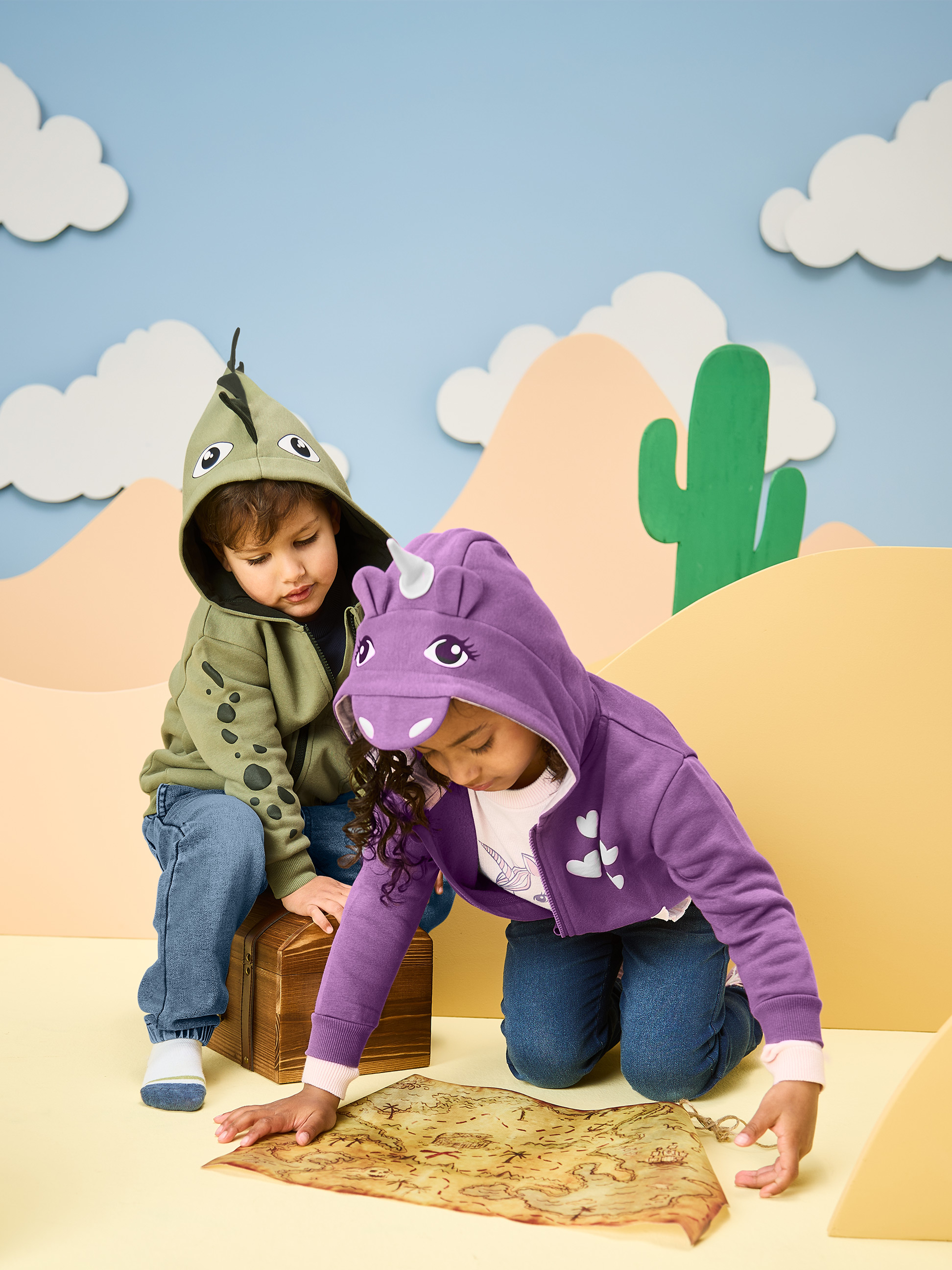 Deti v mikinách s kapucňou dinosaura a jednorožca, pozerajúce na mapu pokladu.
