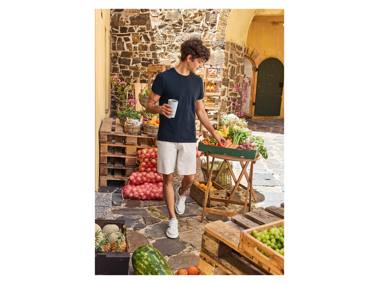 Muž v tričku a šortkách stojí pred stánkom s ovocím.