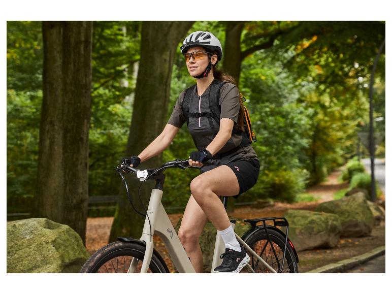 Žena na elektrobicykli s prilbou, okuliarmi a batohom na lesnej ceste.