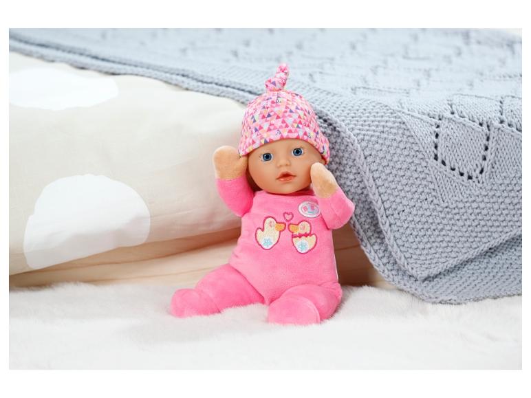 Bábika Baby Born s ružovým oblečením a čiapkou na bielom plyšovom koberci.