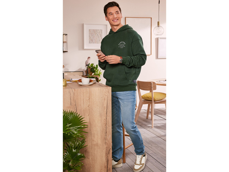 Muž v zelenej mikine s kapucňou s nápisom 'Purpose Fuels' a modrých džínsoch.